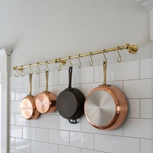 May include: A brass pot rack with several copper and cast iron pans hanging from it. The pans vary in size and shape, with copper exteriors and shiny interiors. The rack is mounted on a white tiled wall, creating a classic kitchen aesthetic.