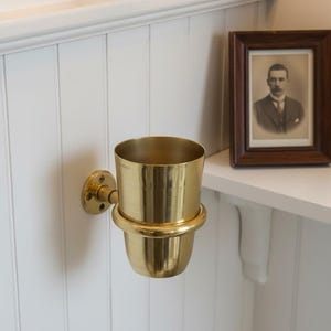 May include: A brass wall-mounted cup holder with a cylindrical design. The holder is attached to a round base with a small, decorative flange. The cup holder is mounted on a white, paneled wall. A framed portrait is visible on a shelf.