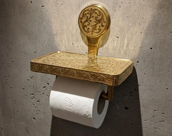 May include: Ornate gold-colored toilet paper holder with a decorative floral pattern. The holder includes a small shelf and a round backplate. A roll of white toilet paper is in place, ready for use. The fixture is mounted on a gray concrete wall.