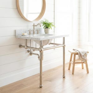 Puede incluir: Un lavabo de baño con encimera de mármol y un soporte de cromo pulido. El lavabo tiene un lavabo blanco y un grifo cromado. Un pequeño taburete de madera con una toalla está al lado del lavabo. Un espejo redondo cuelga de la pared.