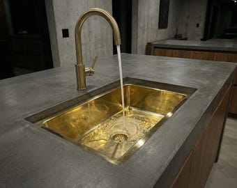 May include: A modern kitchen sink with a gold-toned faucet and a matching gold-colored sink basin. Water flows from the faucet into the sink, creating ripples. The sink is set into a gray countertop, with wooden cabinetry visible below.