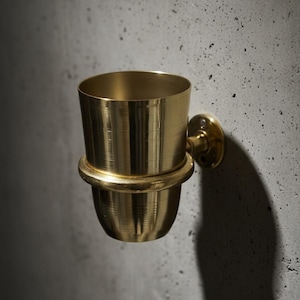 May include: A wall-mounted, brass-colored cup holder with a cylindrical upper section and a rounded lower section. The holder is attached to a circular wall mount. The design features a minimalist aesthetic.