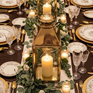 May include: Elegant table setting with gold lanterns, candles, and floral arrangements. The table is set with white plates with gold rims, gold silverware, and crystal glassware. A warm and inviting ambiance for a special occasion.
