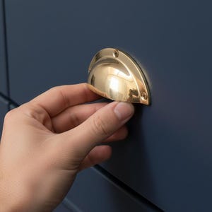 Puede incluir: Un tirador de cajón dorado brillante en forma de copa está montado en una puerta de armario azul oscuro. El tirador está asegurado con tornillos pequeños. La puerta del armario tiene un diseño de panel empotrado.