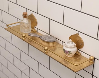 May include: A gold-colored metal shelf with decorative accents, holding a clear glass bottle with a cork stopper, a silver-handled brush, and a small ceramic jar with a floral design. The shelf is mounted on a white tiled wall.