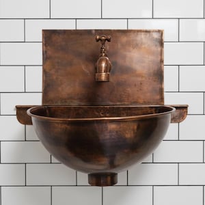May include: A copper-toned wall-mounted sink with a matching faucet. The sink has a rounded basin and a rectangular backplate. The faucet is ornate, and the sink is mounted on a white tiled wall. The sink is a vintage style.