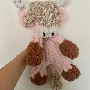 May include: A handmade plush Highland cow in shades of pink, brown, and beige. The cow features a fluffy, textured body, with brown hooves and a shaggy head of fur. The horns are a light gray color, and the cow has black eyes.