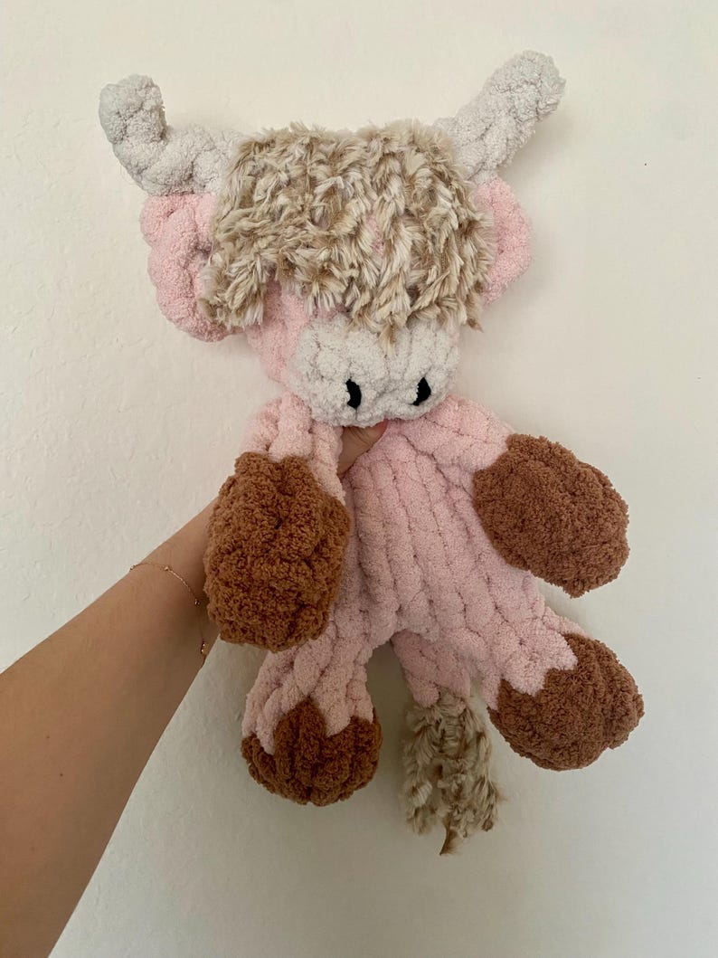 May include: A handmade plush Highland cow in shades of pink, brown, and beige. The cow features a fluffy, textured body, with brown hooves and a shaggy head of fur. The horns are a light gray color, and the cow has black eyes.