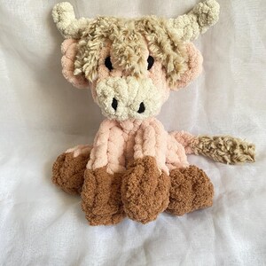 May include: A handmade plush Highland cow in shades of pink, brown, and cream. The soft, cuddly toy features a fluffy head, horns, and hooves. The cow is sitting on a white surface.