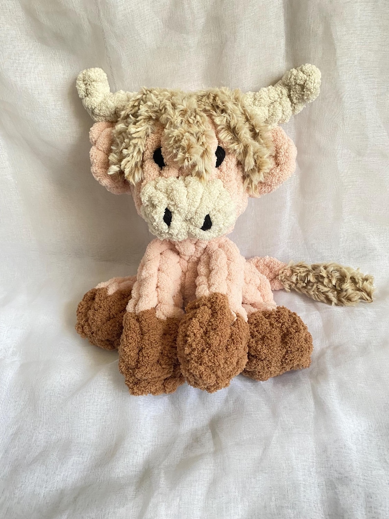 May include: A handmade plush Highland cow in shades of pink, brown, and cream. The soft, cuddly toy features a fluffy head, horns, and hooves. The cow is sitting on a white surface.