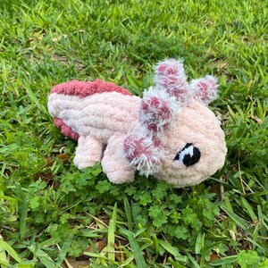 May include: A handmade, plush axolotl toy in shades of pink and burgundy, lying on a bed of green grass. The axolotl features fluffy gills and a detailed face with a black eye. The soft, textured material gives it a cuddly appearance.