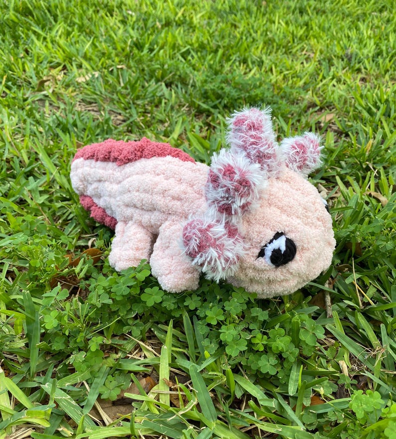 May include: A handmade, plush axolotl toy in shades of pink and burgundy, lying on a bed of green grass. The axolotl features fluffy gills and a detailed face with a black eye. The soft, textured material gives it a cuddly appearance.