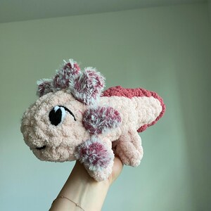 May include: A plush, handmade axolotl toy in shades of pink and white. The soft, cuddly creature features a smiling face with black eyes and decorative frills. The axolotl's body is a light pink, with darker pink accents.