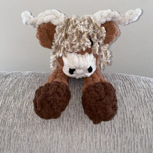 May include: A handmade crochet Highland cow plush toy. The toy features a brown body, legs, and horns, with a white face and fluffy, light brown hair. The cow has black eyes and is made of soft, textured yarn.