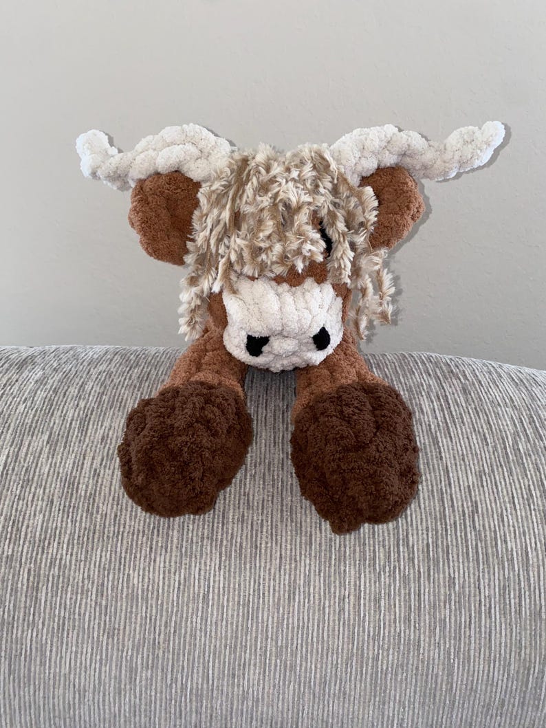 May include: A handmade crochet Highland cow plush toy. The toy features a brown body, legs, and horns, with a white face and fluffy, light brown hair. The cow has black eyes and is made of soft, textured yarn.
