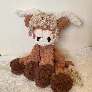 May include: A handmade plush Highland cow toy. The toy features a brown and cream color scheme with a fluffy texture. It has large, dark eyes and long, curved horns. The cow is sitting on a white, textured surface.