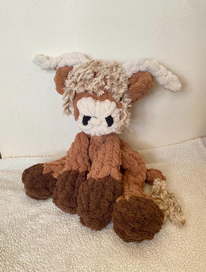 May include: A handmade plush Highland cow toy. The toy features a brown and cream color scheme with a fluffy texture. It has large, dark eyes and long, curved horns. The cow is sitting on a white, textured surface.
