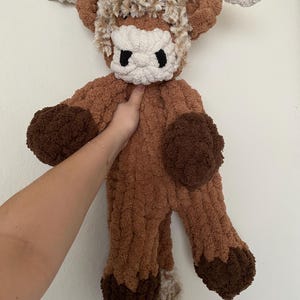 May include: A handmade, plush Highland cow toy in shades of brown, tan, and cream. The cow features a fluffy mane, horns, and a tail. The toy is held up against a white background.