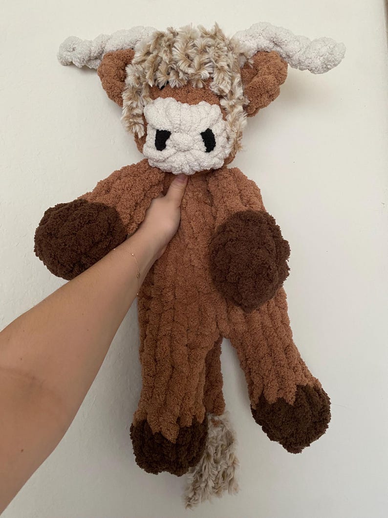 May include: A handmade, plush Highland cow toy in shades of brown, tan, and cream. The cow features a fluffy mane, horns, and a tail. The toy is held up against a white background.