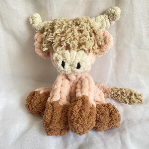 May include: A handmade plush Highland cow in shades of pink, cream, and brown. The cow features a fluffy, textured body, with a cream-colored face, pink ears, and brown hooves. The horns are cream-colored and curled. The toy is soft and cuddly.