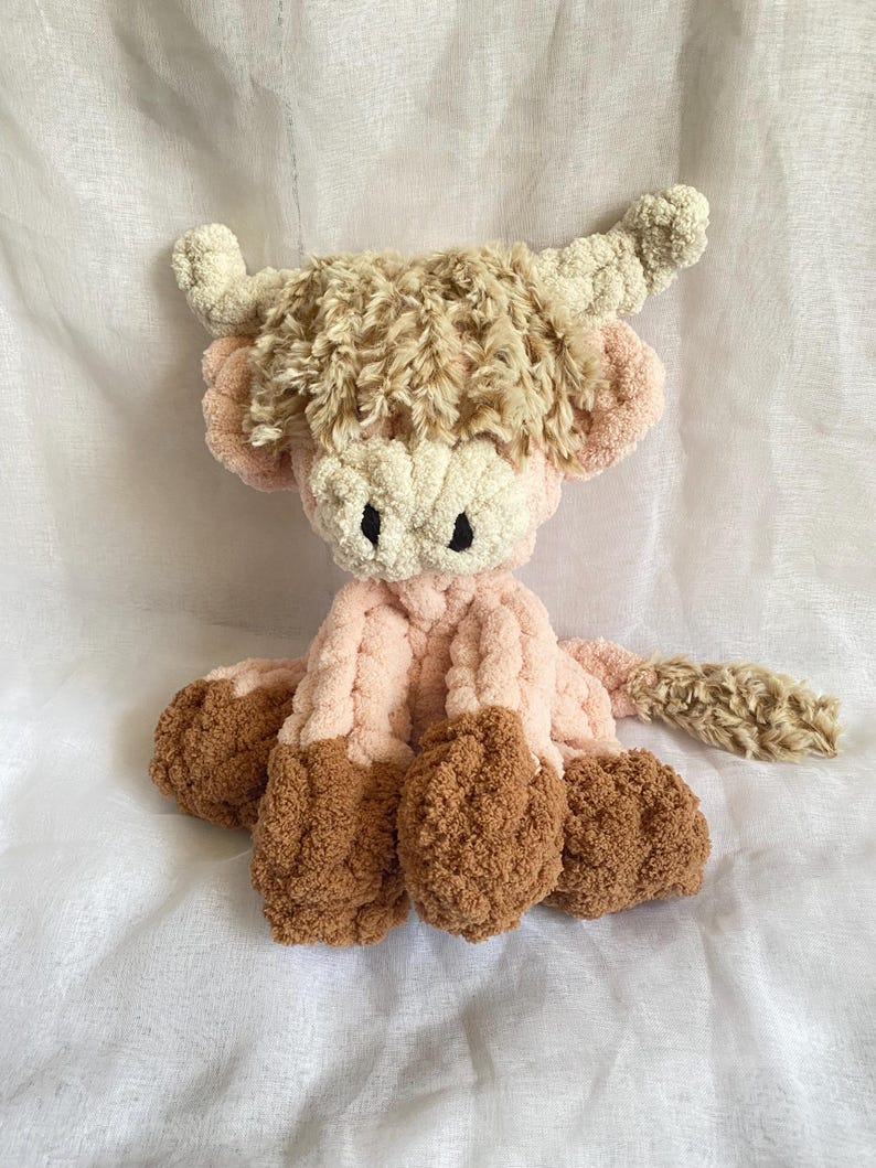 May include: A handmade plush Highland cow in shades of pink, cream, and brown. The cow features a fluffy, textured body, with a cream-colored face, pink ears, and brown hooves. The horns are cream-colored and curled. The toy is soft and cuddly.