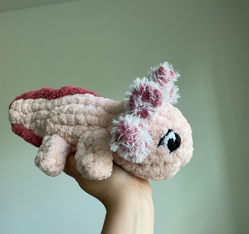 May include: A plush axolotl toy in shades of pink and burgundy. The handmade creature features a textured body, fluffy gills, and a small, black eye. The toy is held in a hand against a neutral background.