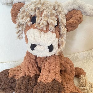 May include: A plush Highland cow stuffed animal in shades of brown, tan, and cream. The toy features a shaggy mane, large horns, and a textured body. The cow's face has black eyes and a white muzzle.