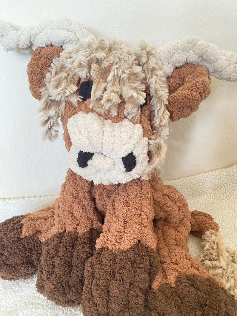May include: A plush Highland cow stuffed animal in shades of brown, tan, and cream. The toy features a shaggy mane, large horns, and a textured body. The cow's face has black eyes and a white muzzle.