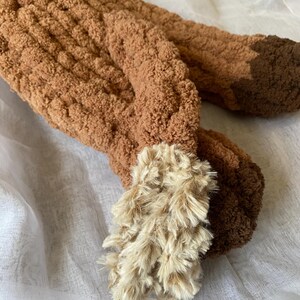 May include: Close-up of a brown, plush, handmade fox tail. The tail is made of a soft, textured material with a lighter, fluffy tip. The tail is lying on a white, textured surface.
