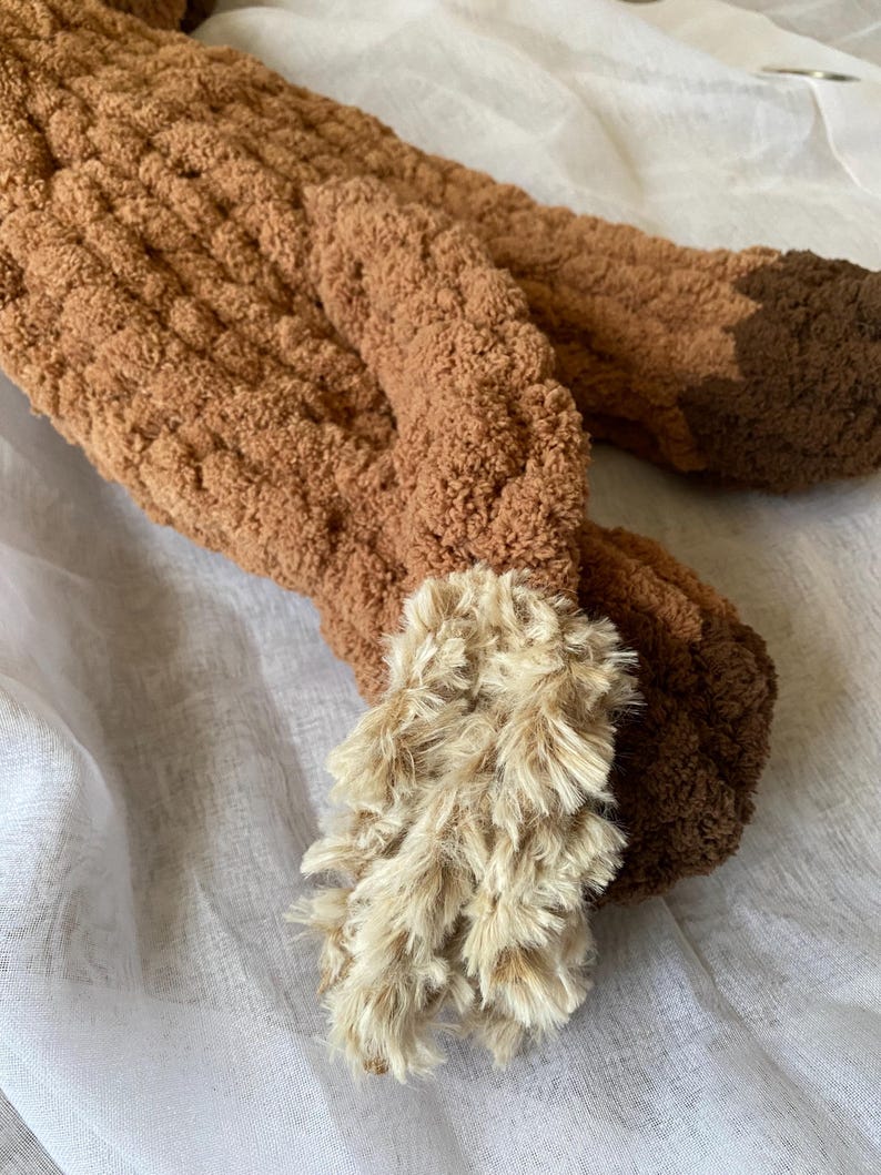 May include: Close-up of a brown, plush, handmade fox tail. The tail is made of a soft, textured material with a lighter, fluffy tip. The tail is lying on a white, textured surface.