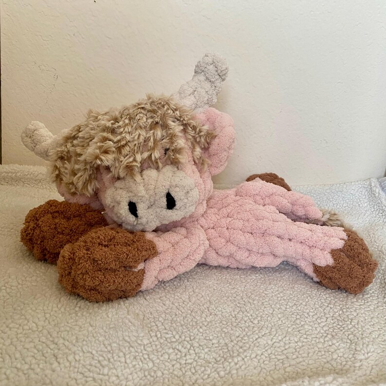 May include: A plush Highland cow in a lying position. The cow features a pink body, brown hooves, and a light brown shaggy mane. The horns are a light gray color, and the eyes are black. The toy is made of a soft, textured material.