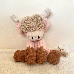 May include: A handmade plush Highland cow in pink, brown, and cream. The cow has a fluffy brown head, pink body, and brown hooves. The horns are a light gray color. The toy is sitting on a white, textured surface.