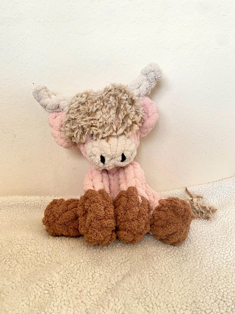 May include: A handmade plush Highland cow in pink, brown, and cream. The cow has a fluffy brown head, pink body, and brown hooves. The horns are a light gray color. The toy is sitting on a white, textured surface.