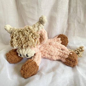 May include: A handmade, plush Highland cow toy in shades of pink, brown, and cream. The soft, cuddly cow has a fluffy head, horns, and feet, with a light pink body. The toy is lying on a white, textured surface.