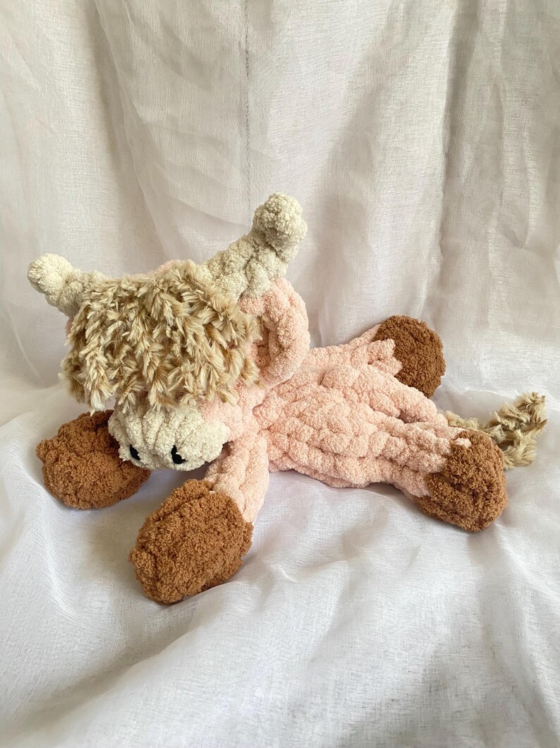 May include: A handmade, plush Highland cow toy in shades of pink, brown, and cream. The soft, cuddly cow has a fluffy head, horns, and feet, with a light pink body. The toy is lying on a white, textured surface.