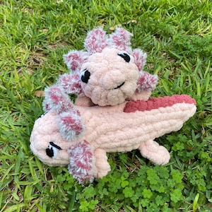 May include: Two handmade crochet axolotl plushies in a grassy setting. The larger axolotl is light pink with a darker pink tail and gills. The smaller axolotl is light pink with darker pink gills. Both have black eyes and are lying on the grass.