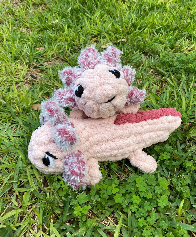 May include: Two handmade crochet axolotl plushies in a grassy setting. The larger axolotl is light pink with a darker pink tail and gills. The smaller axolotl is light pink with darker pink gills. Both have black eyes and are lying on the grass.
