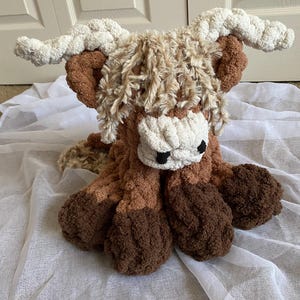 May include: A handmade crochet Highland cow plushie in shades of brown, tan, and cream. The plush toy features a shaggy mane, large horns, and a fluffy body. The cow is sitting on a white fabric.