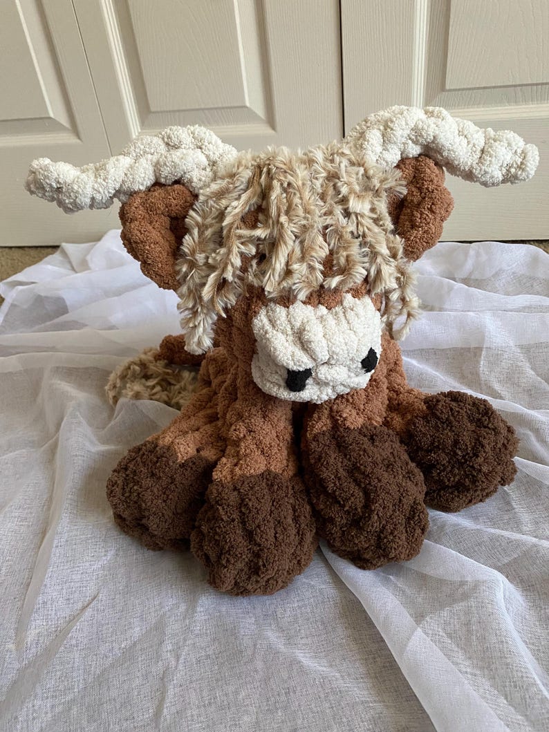 May include: A handmade crochet Highland cow plushie in shades of brown, tan, and cream. The plush toy features a shaggy mane, large horns, and a fluffy body. The cow is sitting on a white fabric.
