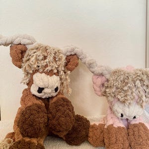 May include: Two plush Highland cow stuffed animals. One is brown and tan, the other is pink and tan. Both have shaggy fur, large horns, and black eyes. The cows are sitting on a white surface.