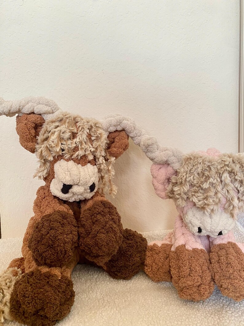 May include: Two plush Highland cow stuffed animals. One is brown and tan, the other is pink and tan. Both have shaggy fur, large horns, and black eyes. The cows are sitting on a white surface.