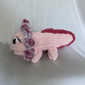May include: A handmade plush axolotl in soft pink with a darker pink trim and accents. The axolotl features fluffy, textured gills in shades of pink and white. The toy is designed with a cute, friendly expression.