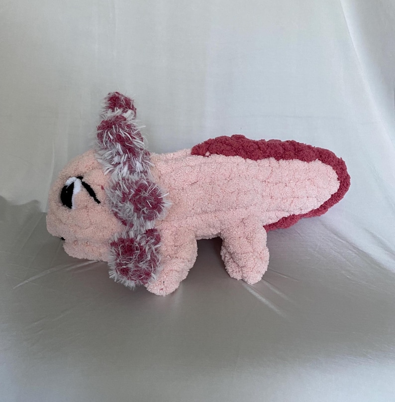 May include: A handmade plush axolotl in soft pink with a darker pink trim and accents. The axolotl features fluffy, textured gills in shades of pink and white. The toy is designed with a cute, friendly expression.
