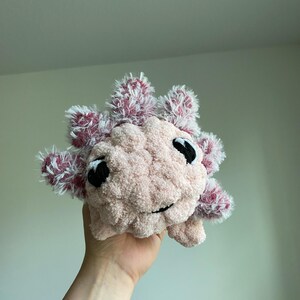 May include: A handmade plush axolotl in soft pink with fluffy, red and white accents. The axolotl has black eyes and a smiling mouth. This cuddly toy is a unique, handcrafted item.