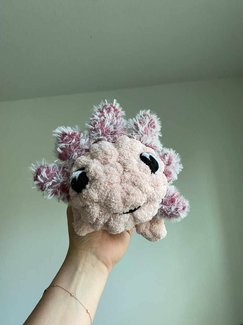 May include: A handmade plush axolotl in soft pink with fluffy, red and white accents. The axolotl has black eyes and a smiling mouth. This cuddly toy is a unique, handcrafted item.