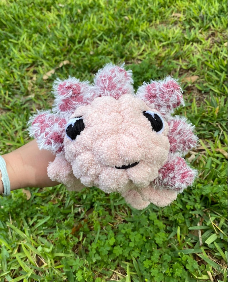 May include: A handmade plush axolotl toy in soft pink with white and pink accents. The axolotl has black eyes and a smiling mouth. The toy is made of a fluffy, textured material and is held against a background of green grass.