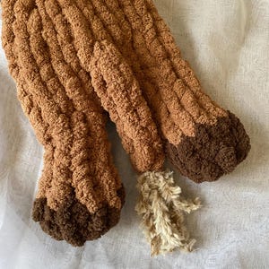 May include: Close-up of a brown, textured, knitted item, possibly a stuffed animal or decorative piece. The item has a light brown body with darker brown accents on the feet and a fluffy, beige tail.