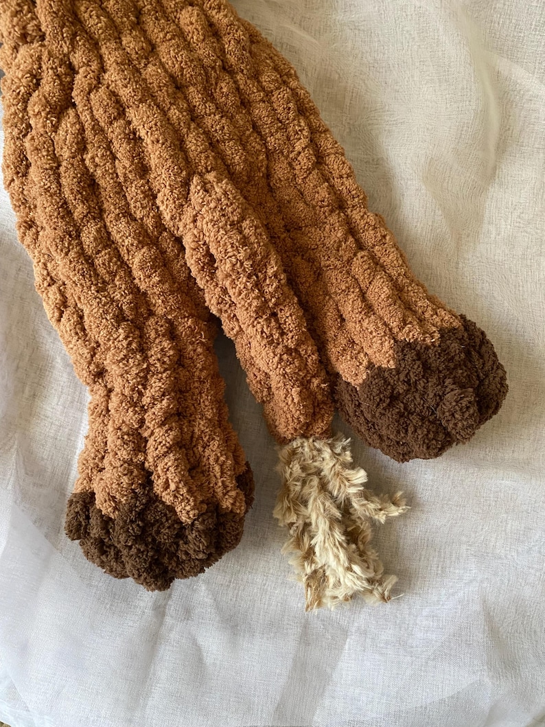 May include: Close-up of a brown, textured, knitted item, possibly a stuffed animal or decorative piece. The item has a light brown body with darker brown accents on the feet and a fluffy, beige tail.