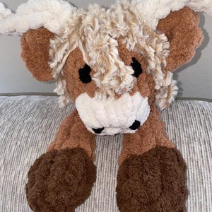 May include: A plush Highland cow toy with a brown body, cream-colored horns, and a fluffy, textured appearance. The toy has black eyes and a white muzzle. The cow's legs are also brown and textured, giving it a soft, cuddly look.