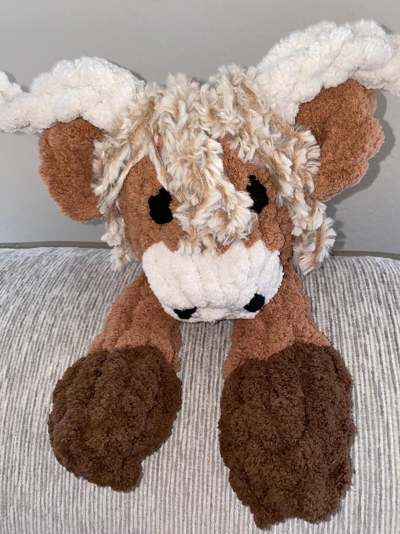 May include: A plush Highland cow toy with a brown body, cream-colored horns, and a fluffy, textured appearance. The toy has black eyes and a white muzzle. The cow's legs are also brown and textured, giving it a soft, cuddly look.